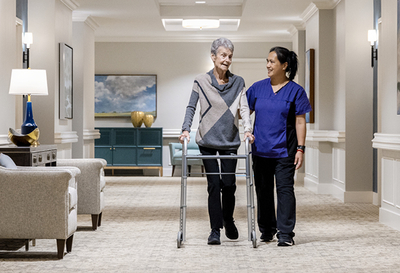 A senior users her walker while a staff in uniform walks alongside her.