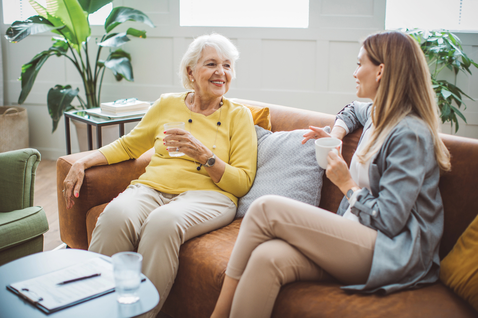 A senior holds a drink while sitting on a couch talking to another woman.