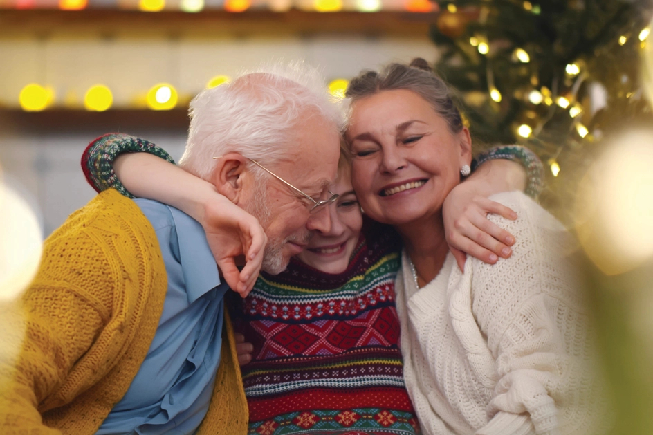 Child hugs seniors on either side of him. A Christmas tree glows in the background.