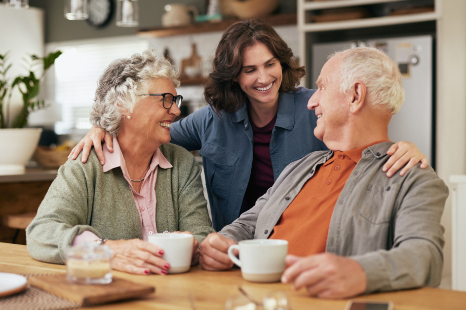 A woman with her arms around a senior couple drinking coffee seated at a table.