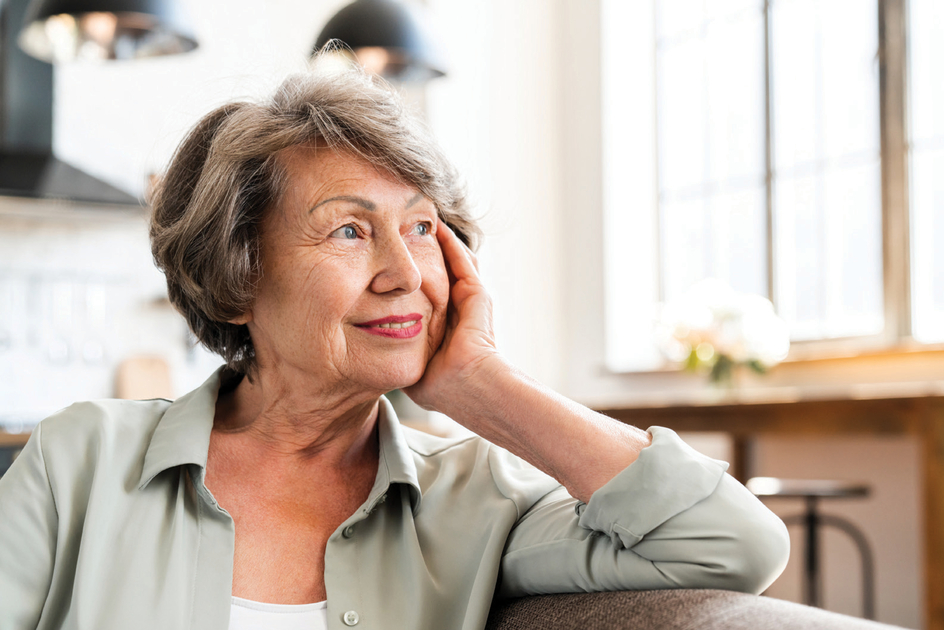Senior woman smiles, putting her hand on her cheek as she sits in a sunny room.