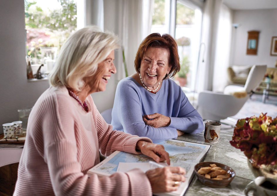 Two senior woman sit at a table, with a book open, laughing together.