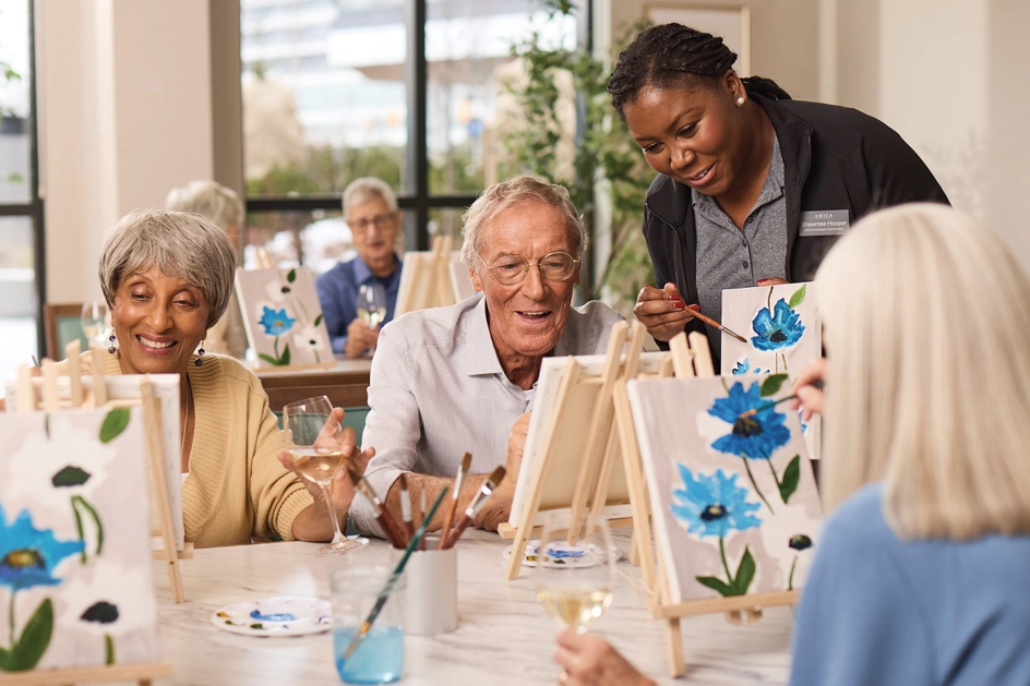 Seniors paint on easels on tables with support from an Amica employee.