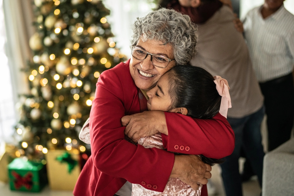 A senior smiles while hugging a child in front of a decorated tree, lit for Christmas.
