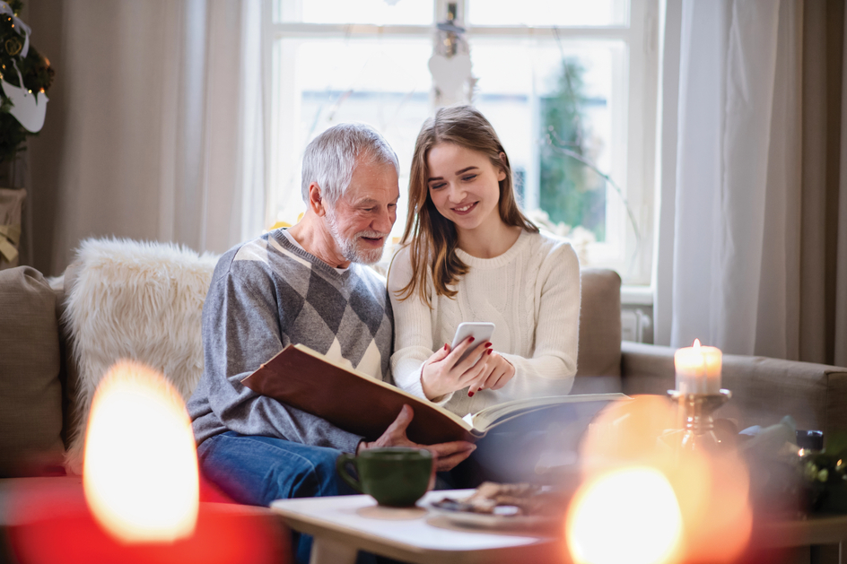 Senior and young woman sit together, looking at smartphone while also holding a photo album