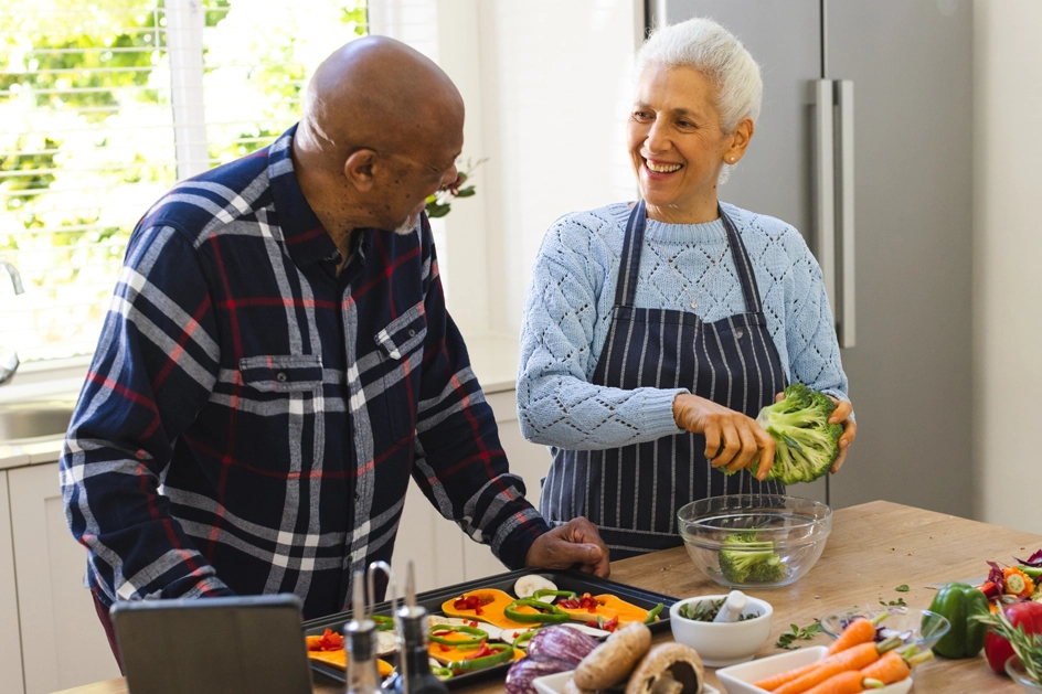 A senior couple cut vegetables up, arranging them on a baking sheet.