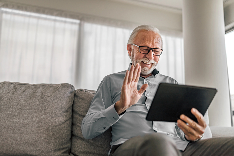 A senior raises his hand to wave while holding up a tablet while sitting on a couch.