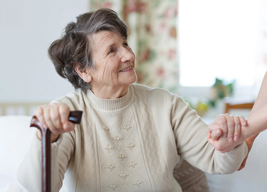 A senior woman sits, holding a cane. She's smiling, looking up to someone standing, who's holding her hand.