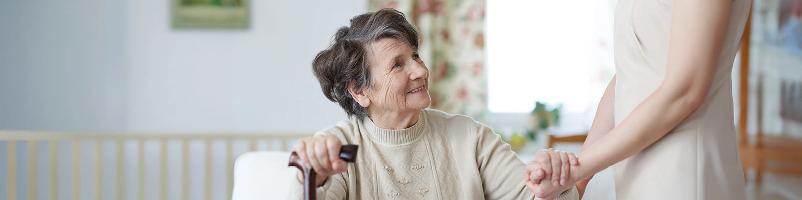 A senior woman sits, holding a cane. She's smiling, looking up to someone standing, who's holding her hand.