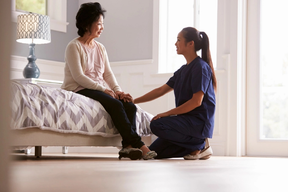 A woman kneels next to a senior woman who's sitting on a bed.
