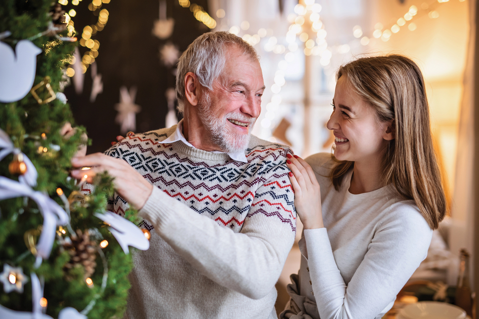 A senior and younger woman smile as they stand by the Christmas tree.