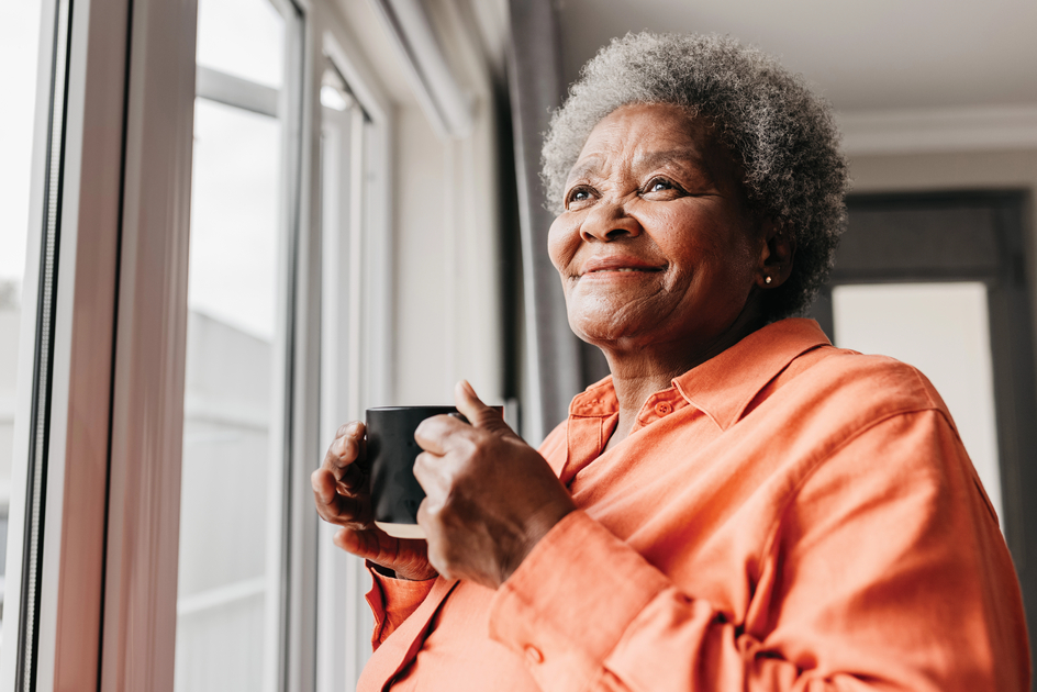 A woman stands, looking out the window while holding a mug.