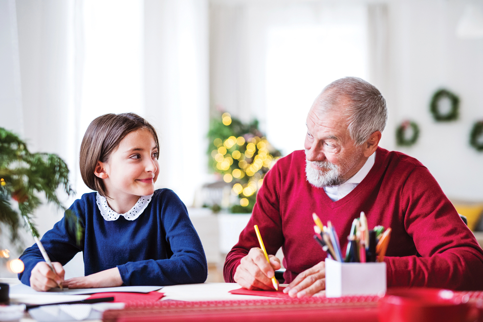 Girl and senior, dressed in festive colours, sit together drawing on a table with a red tablecloth