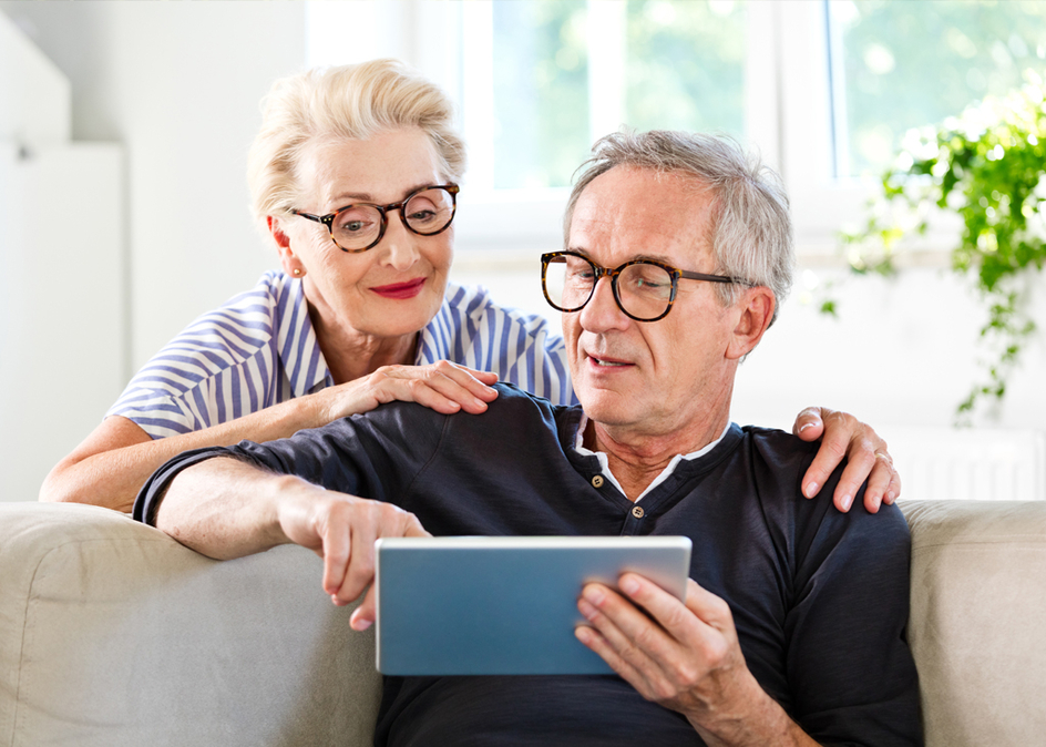 Senior man sitting on sofa in the living room at home and showing something on digital tablet his wife. Senior woman peeking on screen.