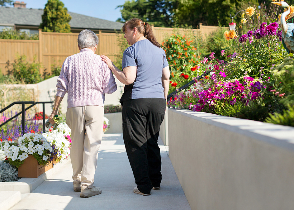Senior and caregiver walking through a garden outside.
