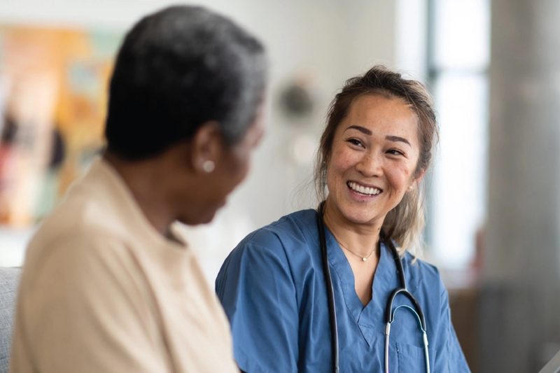A woman in scrubs, and with a stethoscope, smiles while talking to a senior beside her.