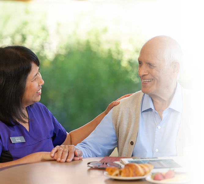 Amica employee sits, smiling, with senior at table.