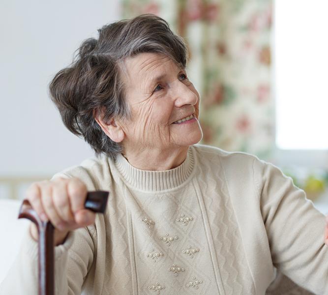 A senior woman sits, holding a cane. She's smiling, looking up to someone standing, who's holding her hand.