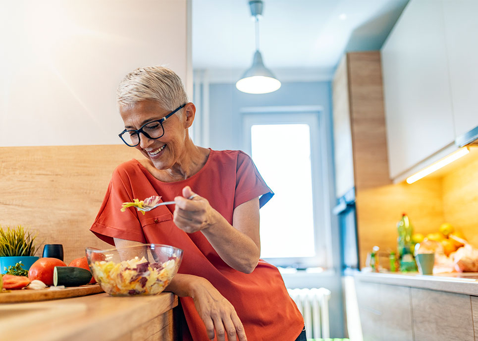 Senior woman smiling and standing in the kitchen while eating a salad.