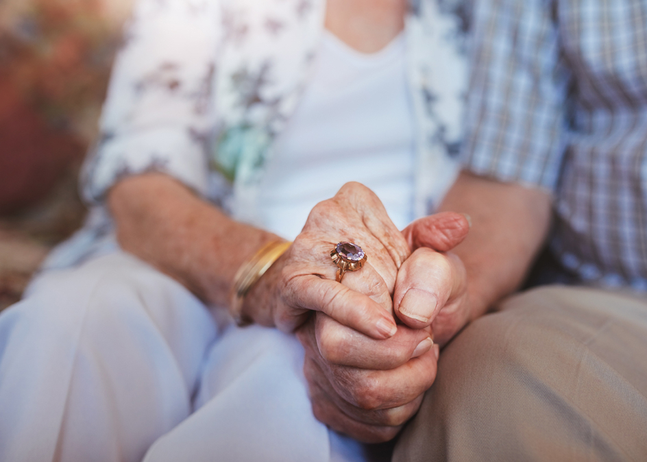 Cropped shot of elderly couple holding hands while sitting together at home. Focus on hands.