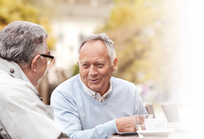 Two senior men sit at a table, chatting while drinking water and tea.