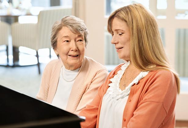 Senior watches as woman plays piano beside her.