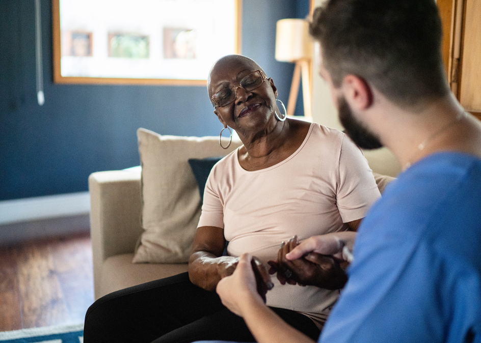 A senior woman closing her eyes, holding hands with a caregiver on a couch in a living room.