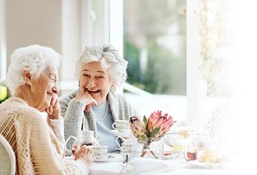 Two women sit laughing together over a cup of tea while sitting by a window.