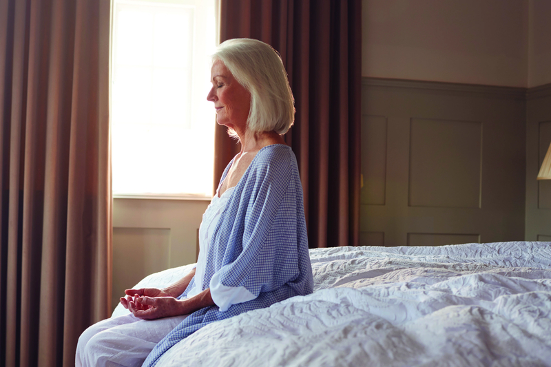 A senior woman, in a nightgown and robe, sits at the end of her bed meditating by a window.