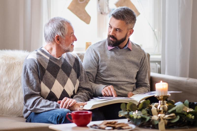 Man sits on a couch with a senior man. They are looking at a photo album while having a hot drink.