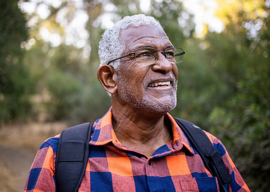 A close-up of a senior man hiking outside in the woods wearing a plaid button up shirt and backpack.