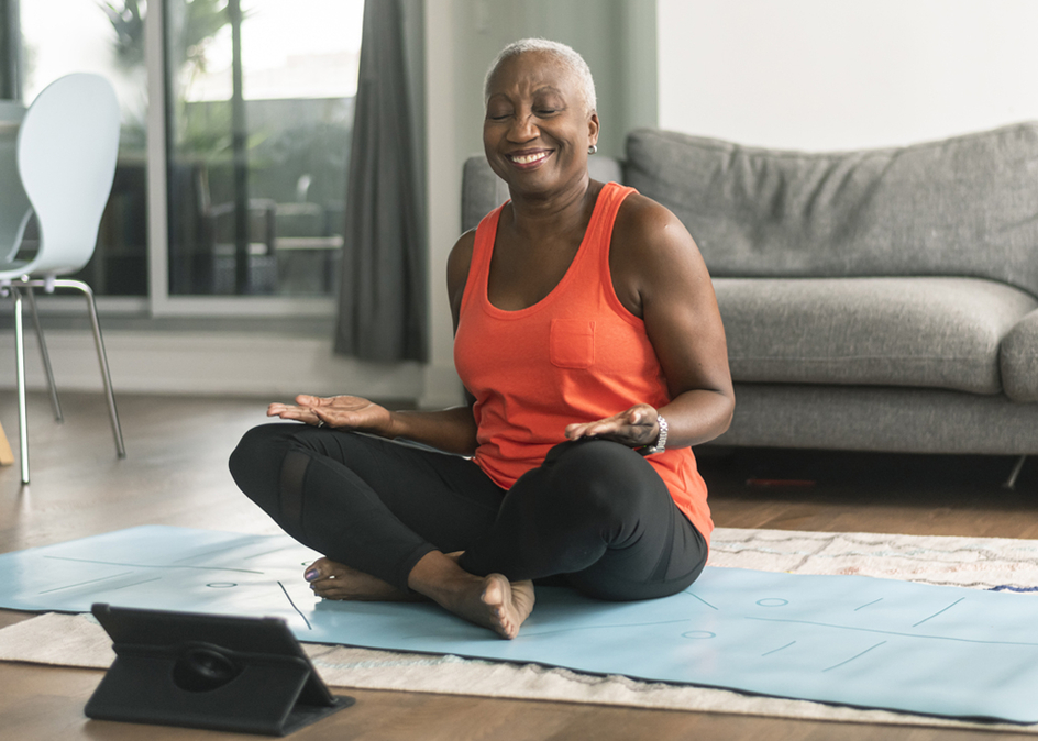 A senior woman takes an online yoga class. She is interacting with the teacher on her tablet and about to commence her class. She is wearing casual active wear and is taking the class in her lounge room. She is sitting on a blue yoga mat.
