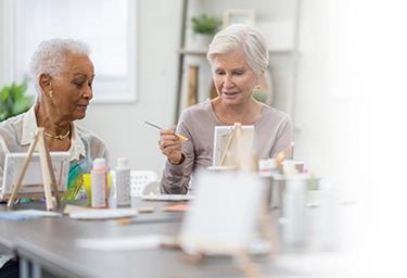Senior women paint together on small easels at a table.