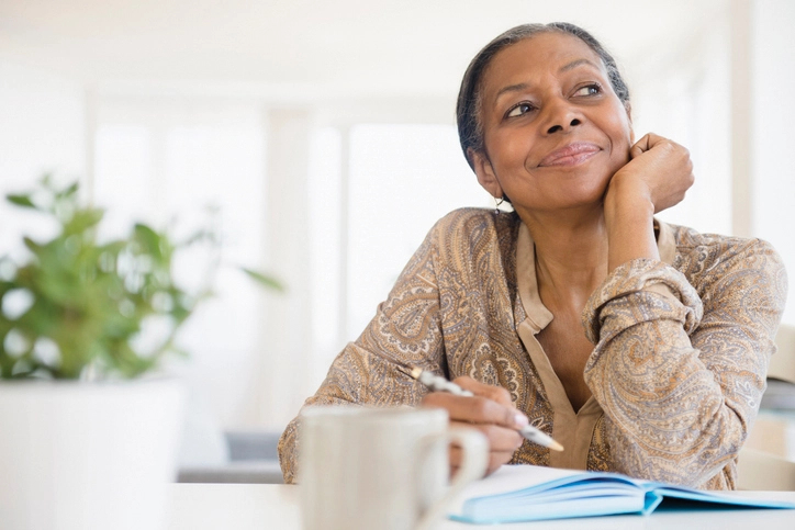 Senior woman sits holding a pen as she sits at a desk and looks up, thinking.