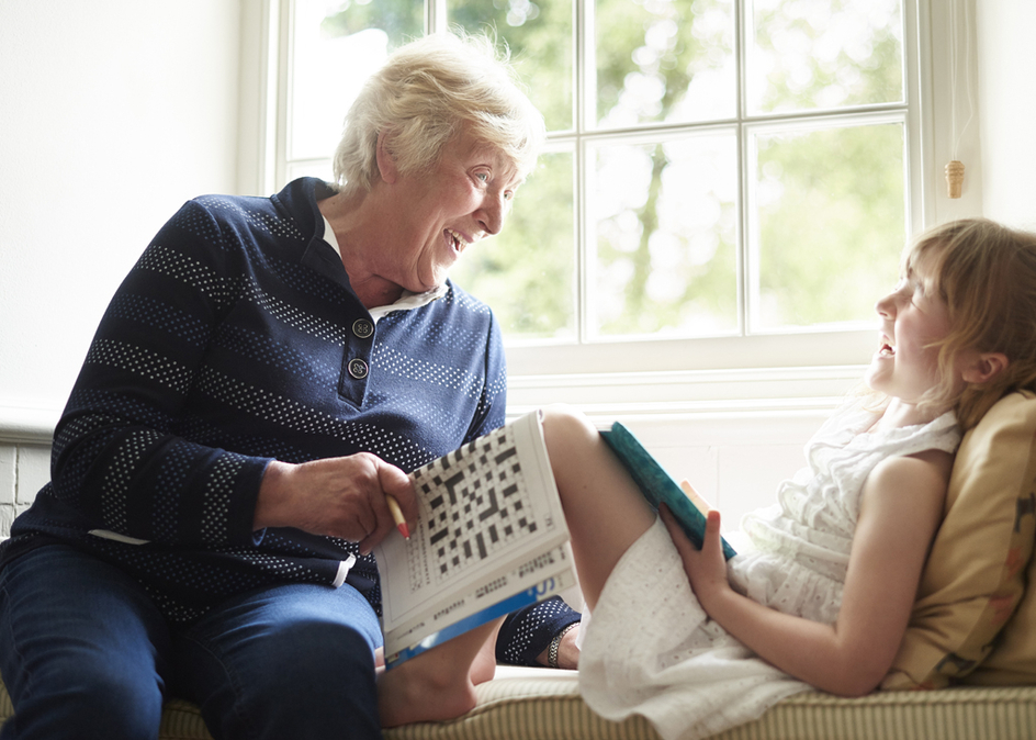 A little girl and her grandmother are sitting in a window seat of a house both interacting with their chosen media. The grandmother is filling out a crossword puzzle whilst the little girl plays on her digital tablet.