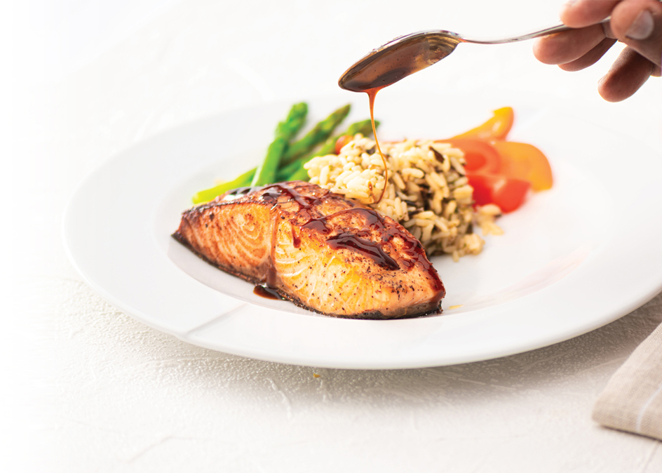 A chef's hand spooning honey ginger glaze over a plate of pan seared salmon