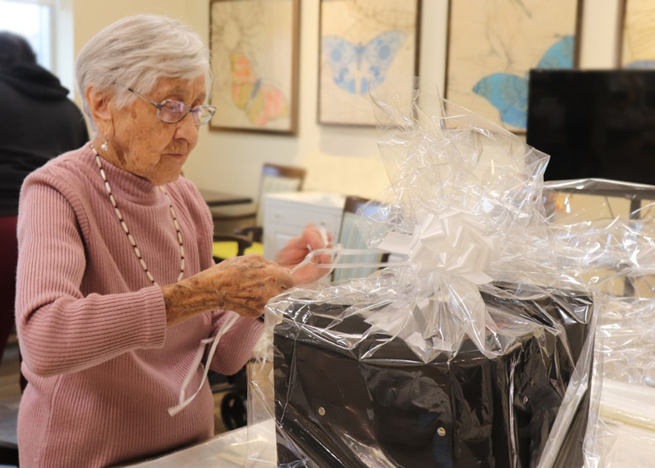 A senior woman in a pink sweater wrapping a basket with clear cellophane and a large white ribbon.