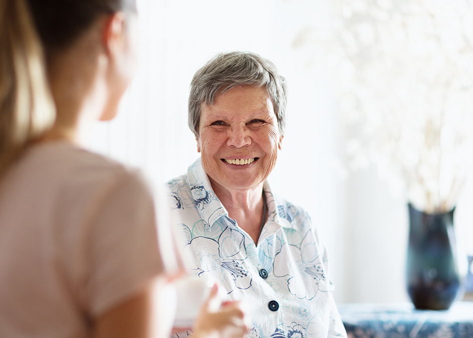 Young woman chatting with senior woman, indoors in living room, sitting on sofa. They are having a cup of coffee, smiling at each other