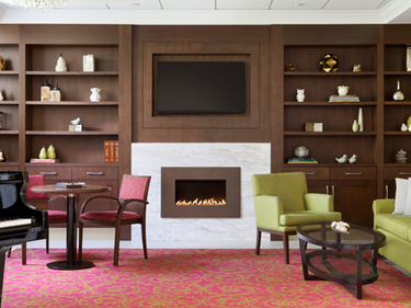 Seating area at Amica Unionville's Bistro: at the back there is a dark wooden closet with decorative elements on its shelves, a TV and a fireplace, and the room is furnished with couches, armchairs and a piano.