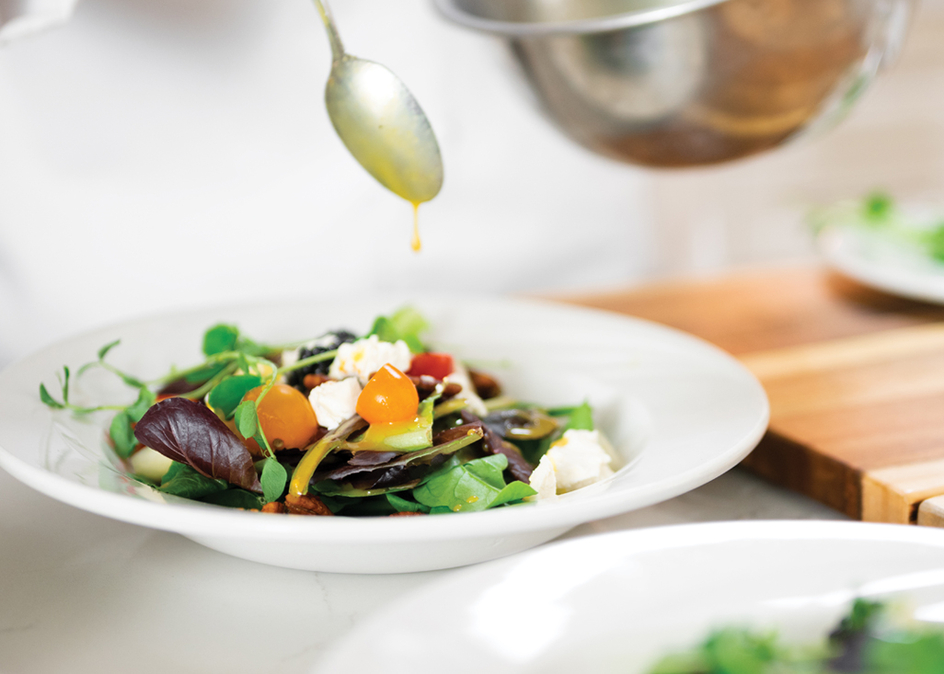 A chef's hand spooning honey vinaigrette dressing over a bowl of salad