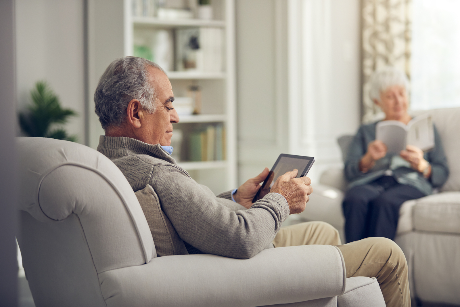 Male resident sitting in chair with iPad, across from female resident reading book on couch