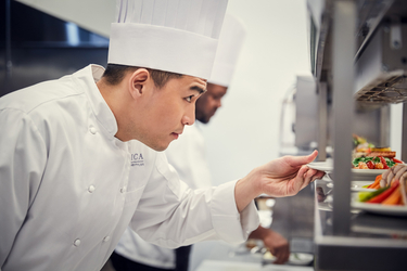 Amica Chef placing a plate of food on the pass in the back-of-house kitchen